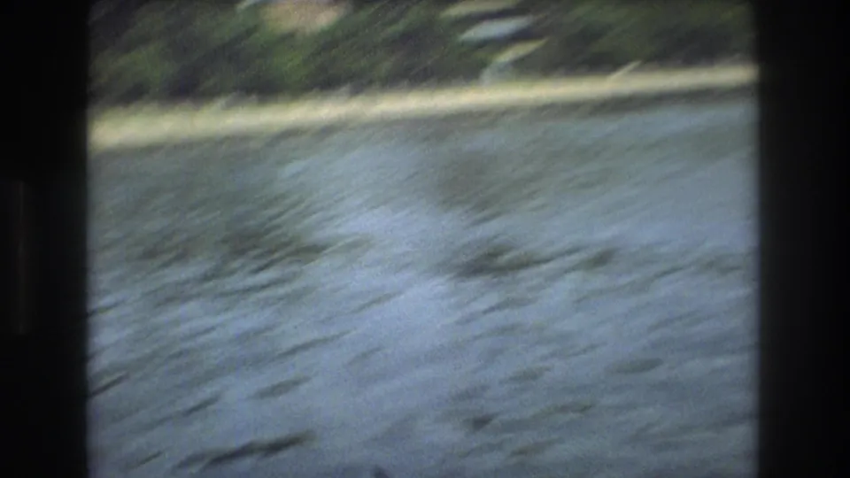 Motion-blurred water fills the frame with a green shoreline at the top, suggesting a moving boat perspective in archival 8mm footage.