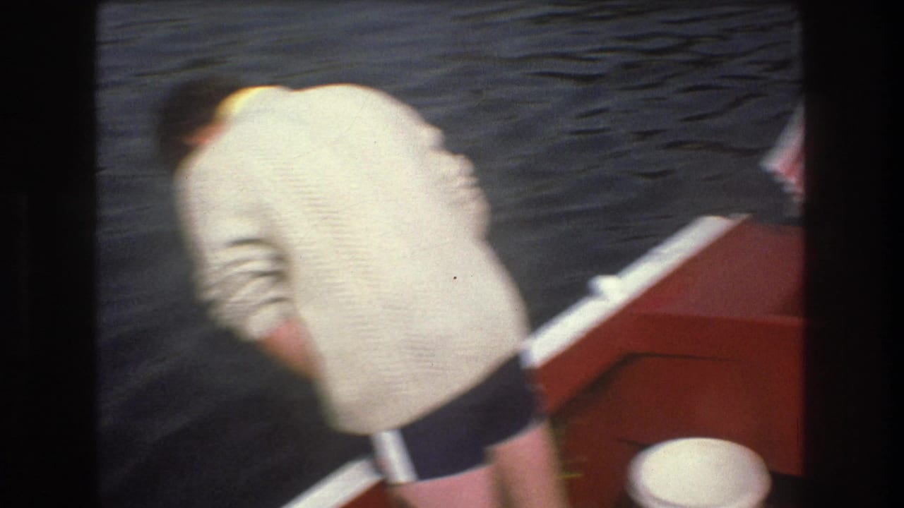 A person in a light sweater and dark shorts leans near the side of a red boat, with dark water behind them in the 1975 Boston home-movie clip.