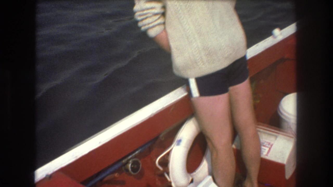 A person in a cream-colored sweater and dark shorts stands at the stern of a red boat beside a white life preserver, rope, and bucket in 1975 Boston boating footage.