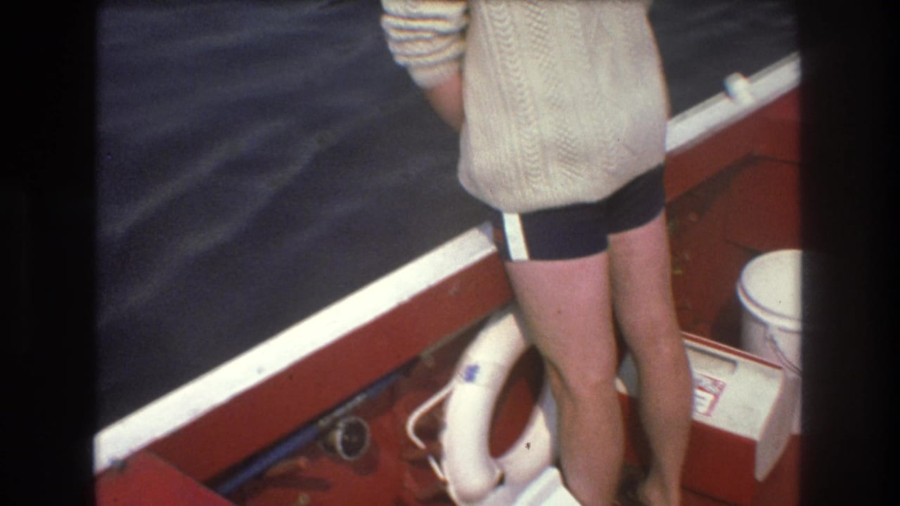 1975 Boston-area 8mm frame showing a person from behind in a cream cable-knit sweater and dark striped trunks standing near the edge of a red boat beside choppy water.