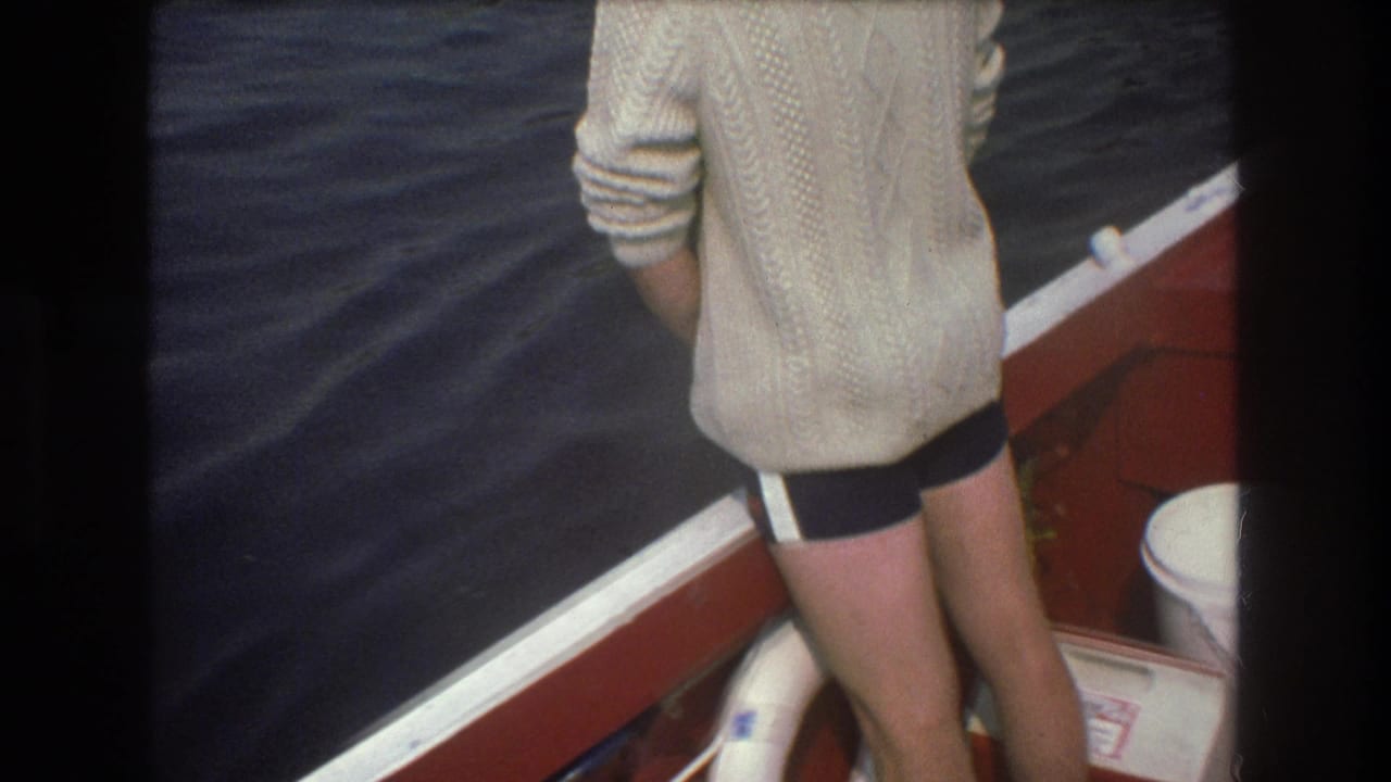 1975 Boston-area 8mm frame with a rear view of a person in a cream sweater standing by white boat trim as dark water passes alongside.