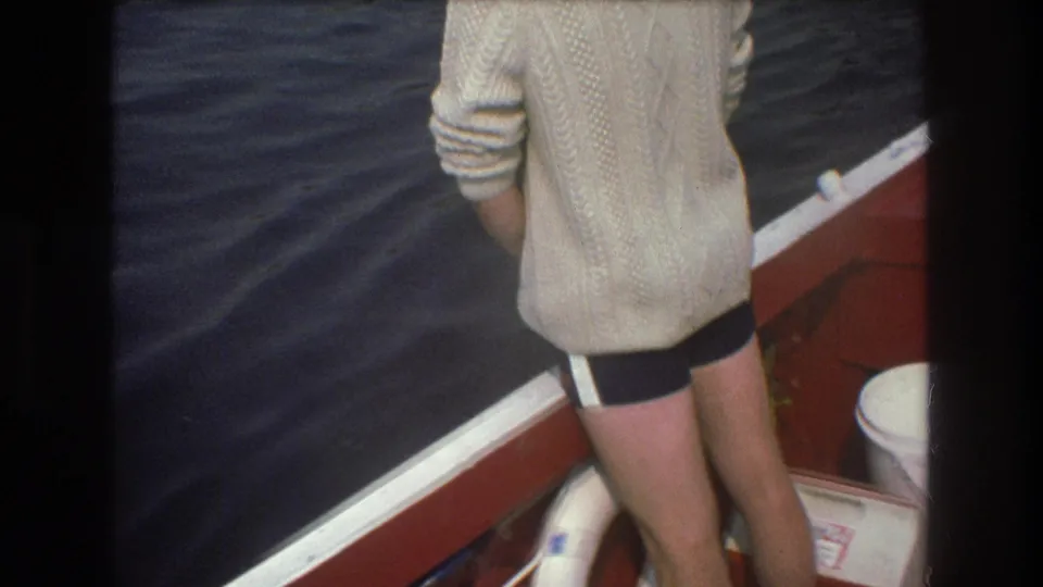 1975 Boston-area 8mm frame with a rear view of a person in a cream sweater standing by white boat trim as dark water passes alongside.