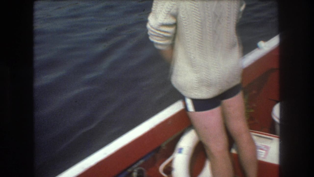 1975 Boston-area home-movie frame showing a person from behind on a red boat deck, with a white life preserver below and dark moving water beyond the rail.