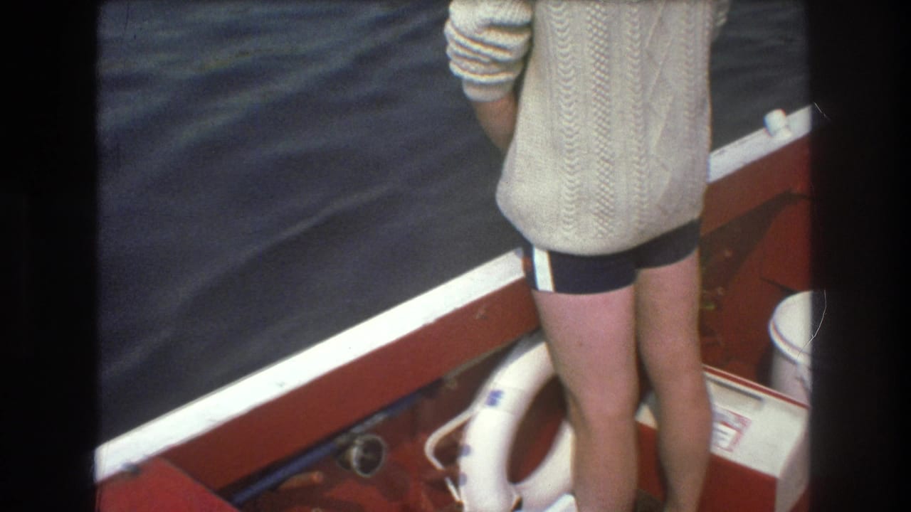 1975 Boston-area 8mm frame showing a person in a cream sweater and dark trunks standing by the white-trimmed edge of a red boat as choppy water fills the background.