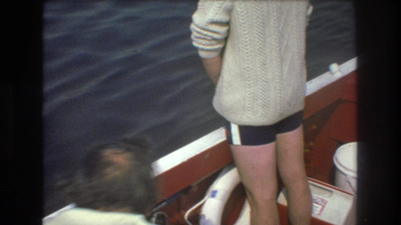 1975 Boston home-movie frame showing a person in a cream sweater standing by the red boat edge while another blurred person bends into the lower left of the shot.