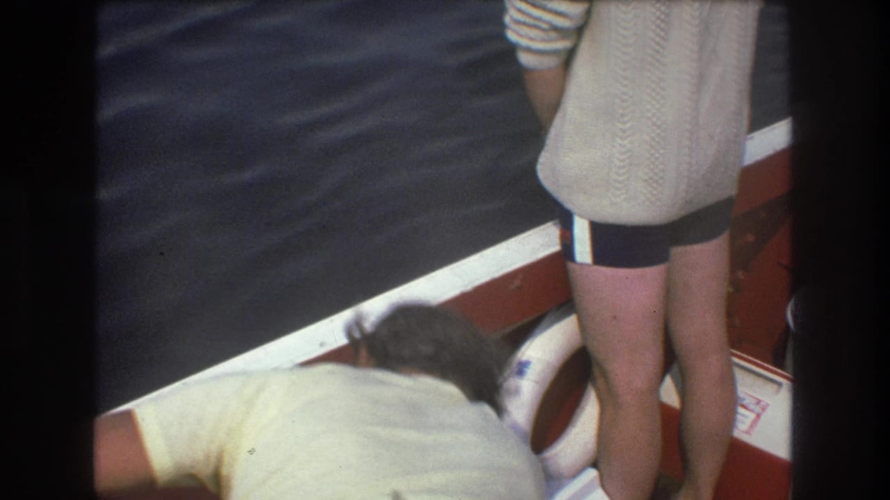 1975 Boston home-movie frame with one adult man standing at the stern and another leaning forward near the side of the boat, with a white life preserver and rippling water visible.