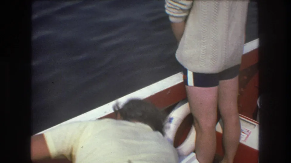 1975 Boston home-movie frame with one adult man standing at the stern and another leaning forward near the side of the boat, with a white life preserver and rippling water visible.