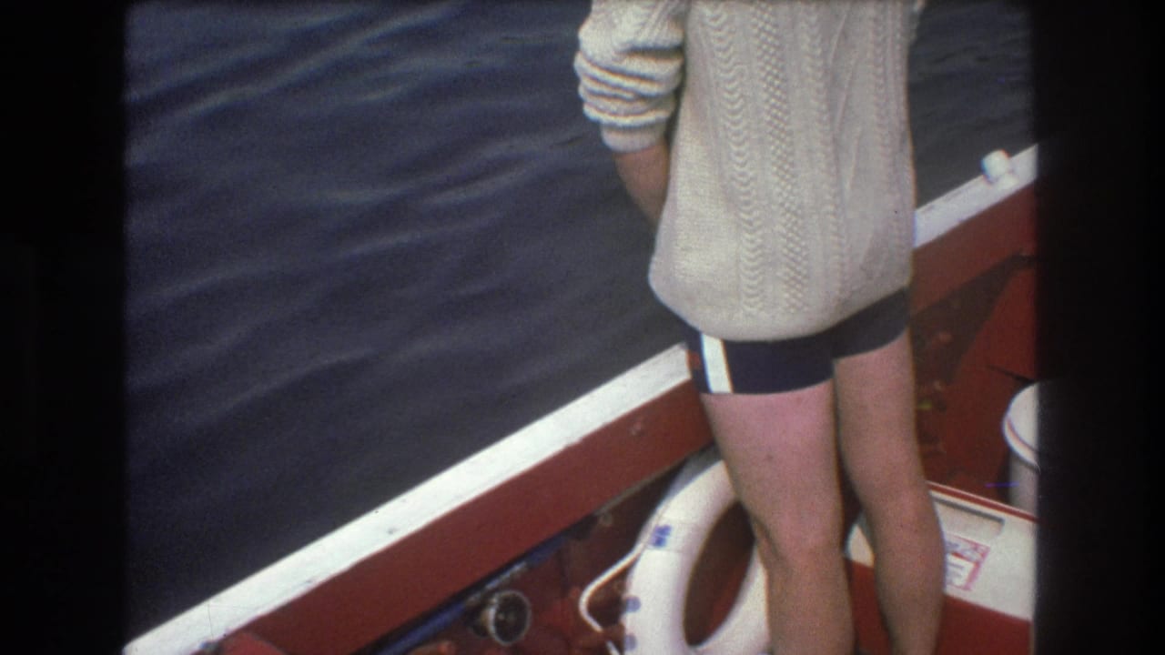 1975 Boston home-movie frame showing the standing man in a cable-knit sweater and striped shorts near the stern of a red-and-white boat, with dark water and a life preserver in view.