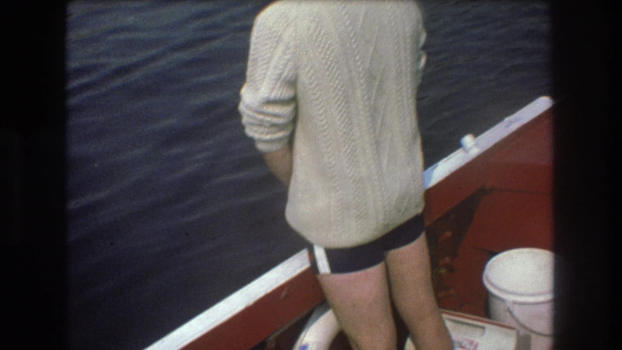 1975 Boston home-movie frame focused on an adult man standing at the stern of a red-and-white boat, with a white container, rail trim, and dark rippling water nearby.