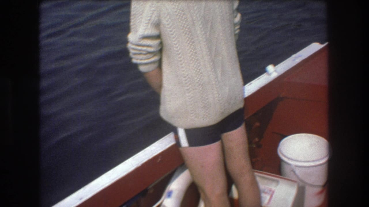 In metadata-supported 1975 Boston 8mm footage, a man in a cream cable-knit sweater and dark shorts stands with his back to the camera at the side of a red boat over rippling water.