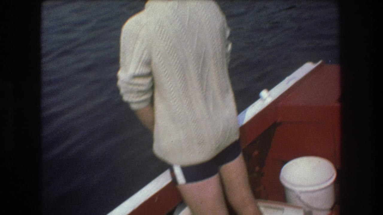 Man stands with his back to the camera beside the red boat rail and water, with a white bucket nearby.