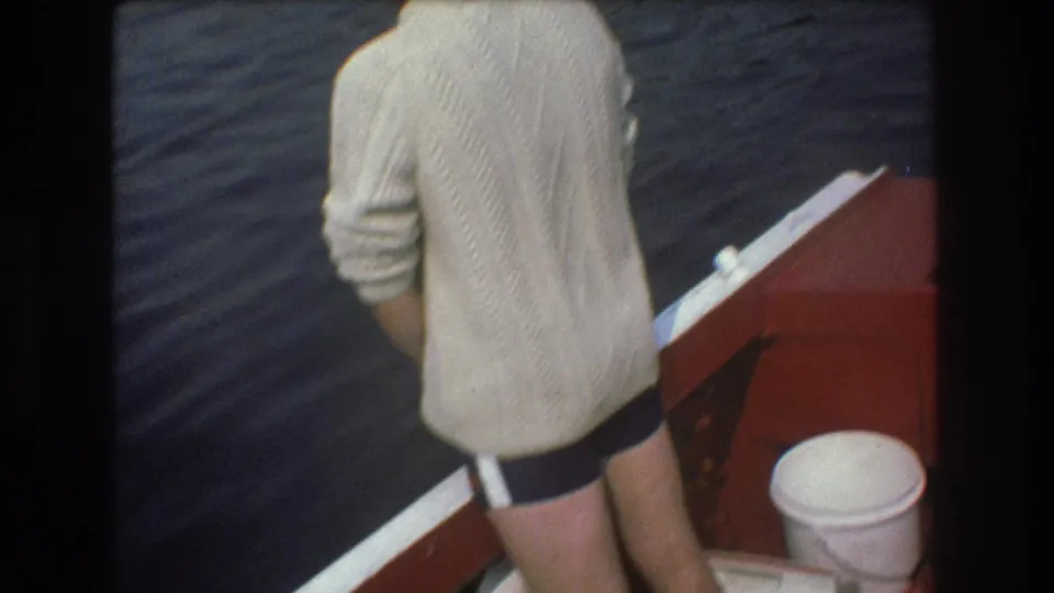 The man remains at the edge of the red boat in the metadata-supported 1975 Boston home-movie footage, facing the water with a white bucket visible on deck.