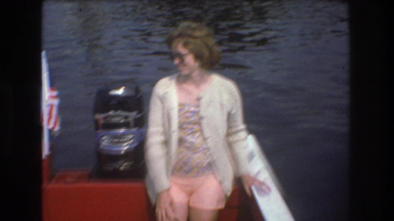 A woman in sunglasses, a patterned dress, and a light cardigan stands on the red boat near a black outboard motor in metadata-supported 1975 Boston home-movie footage.