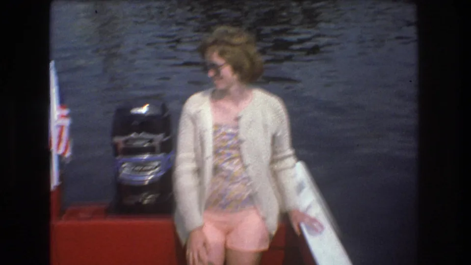 A woman in sunglasses, a patterned dress, and a light cardigan stands on the red boat near a black outboard motor in metadata-supported 1975 Boston home-movie footage.