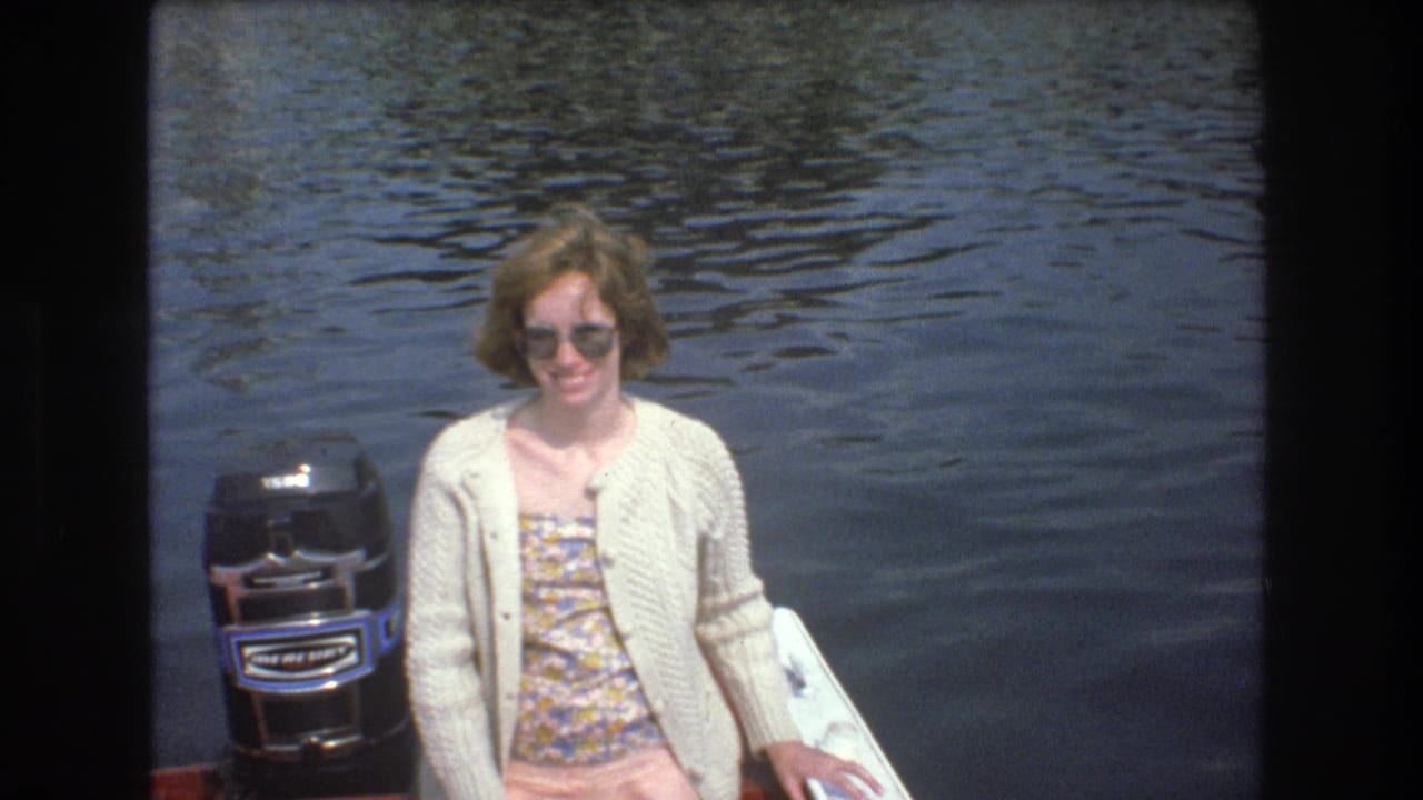 A woman smiles toward the camera on the red boat, wearing sunglasses and a light cardigan beside a black outboard motor, with rippling water behind her in the 1975 Boston home-movie scene.