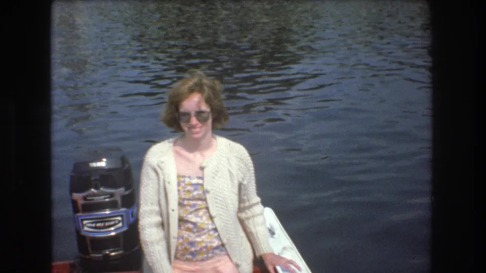 A woman smiles toward the camera on the red boat, wearing sunglasses and a light cardigan beside a black outboard motor, with rippling water behind her in the 1975 Boston home-movie scene.
