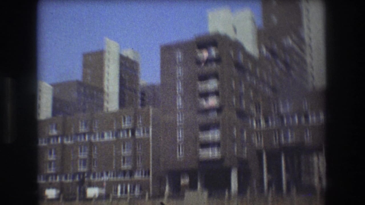Soft 8mm frame of Boston waterfront apartment buildings in 1975, with brick facades, balconies, shoreline edge, and blue sky visible from the water.