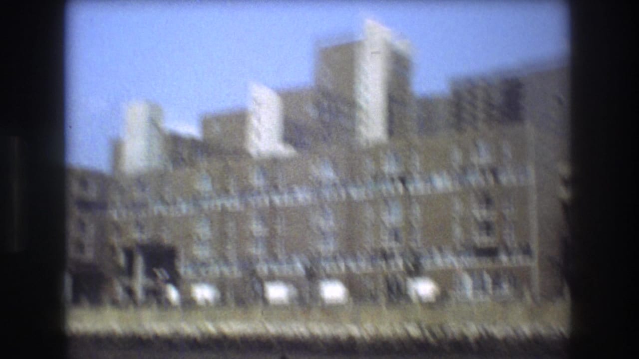 Boston waterfront view from a moving vessel showing brick residential buildings near the shore and taller city structures behind them in soft 8mm focus.