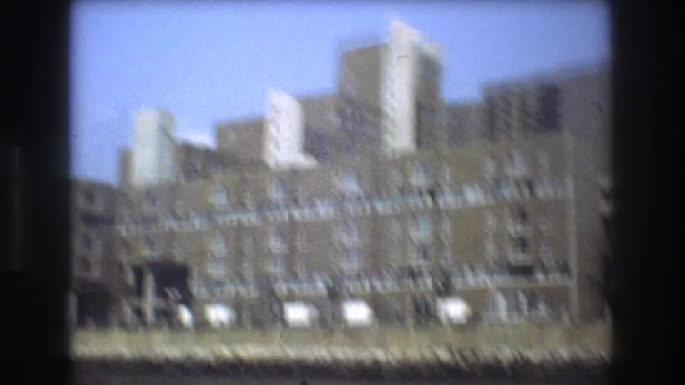 Boston waterfront view from a moving vessel showing brick residential buildings near the shore and taller city structures behind them in soft 8mm focus.