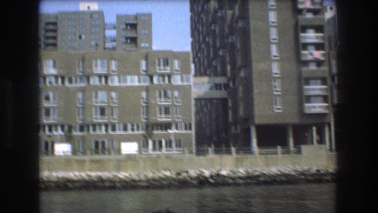 Boston waterfront apartment buildings in a 1975 8mm frame, with balconies, a rocky shoreline or seawall, water in the foreground, and taller structures behind.