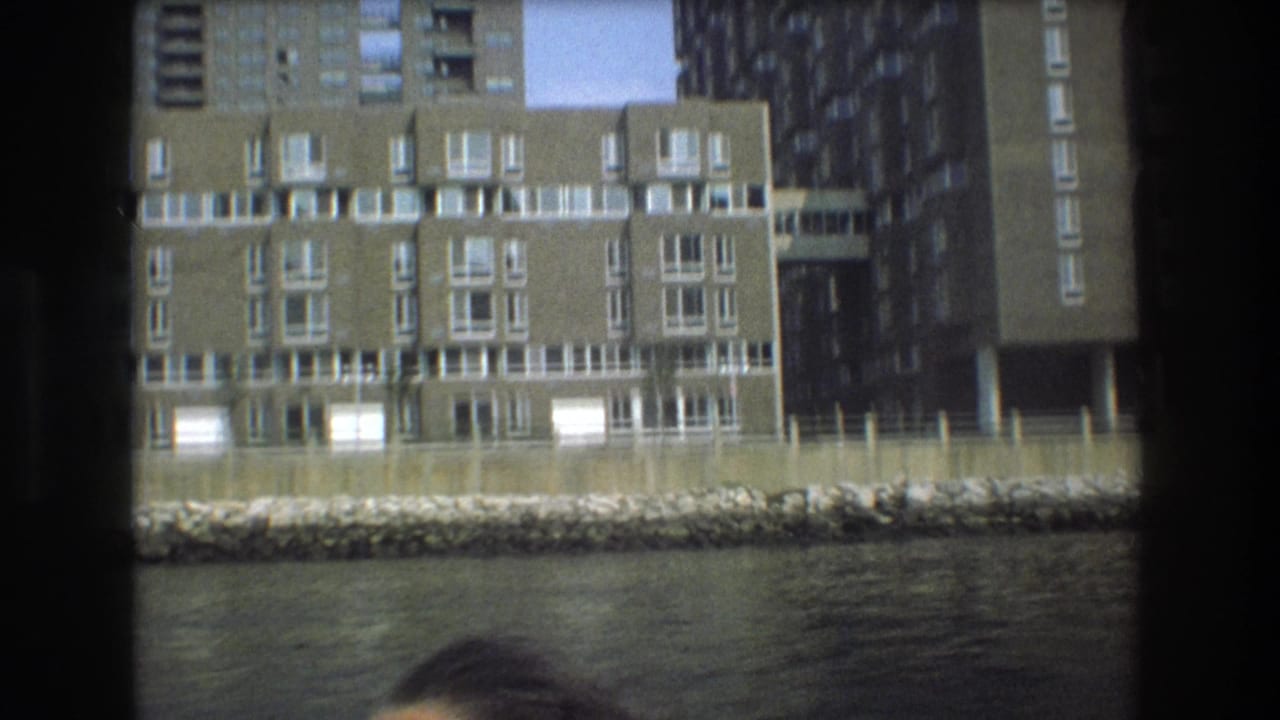 Boston 1975 home-movie frame showing brick and concrete apartment buildings behind a stone shoreline, with water in the foreground and dark boat framing at the sides.