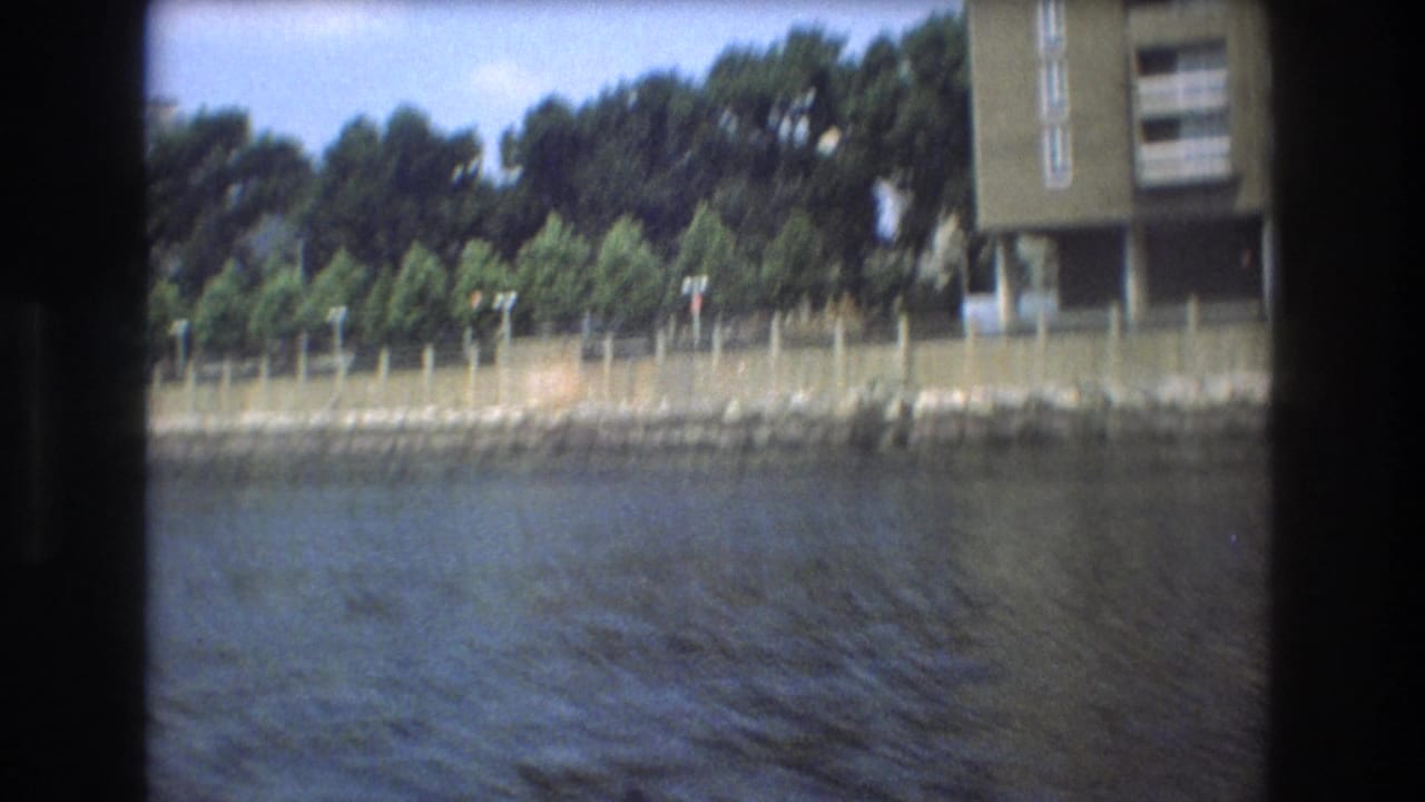 Boston 1975 waterfront view from a moving boat, with trees lining a fenced shoreline and apartment structures partly visible near the frame edges.