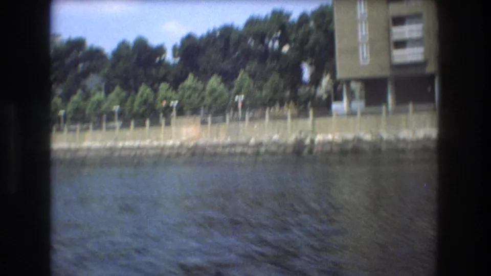 Boston 1975 waterfront view from a moving boat, with trees lining a fenced shoreline and apartment structures partly visible near the frame edges.