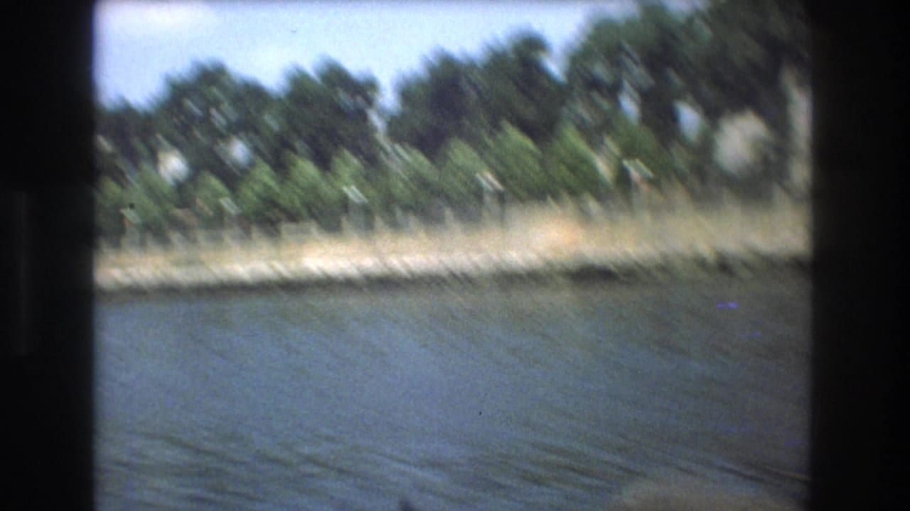 Motion-blurred Boston 1975 home-movie frame of a tree-lined waterfront and low fence seen across rippling water from a moving boat.