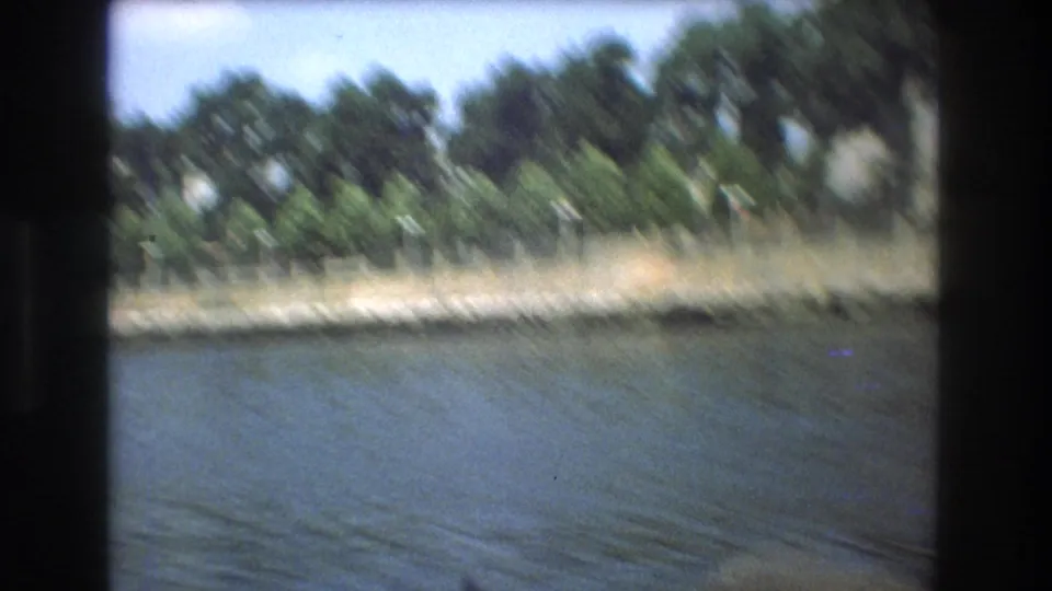 Motion-blurred Boston 1975 home-movie frame of a tree-lined waterfront and low fence seen across rippling water from a moving boat.