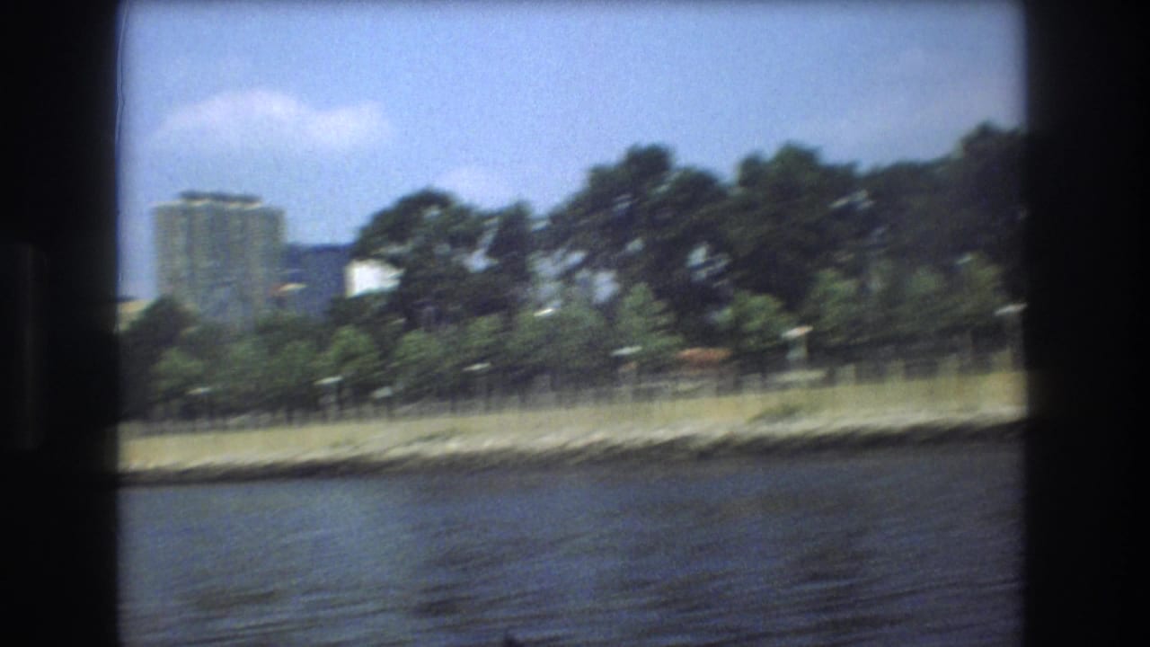 1975 8mm boat view across rippled water toward a Boston shoreline with trees and distant high-rise buildings under a pale sky.