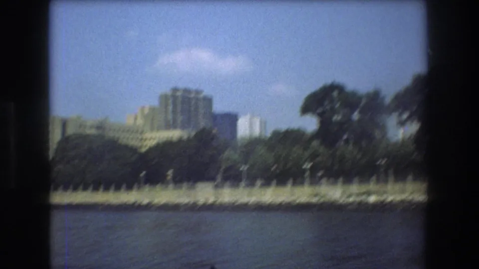 Moving 8mm view from a boat showing Boston waterfront trees, a low shoreline edge, and large buildings set back from the river.