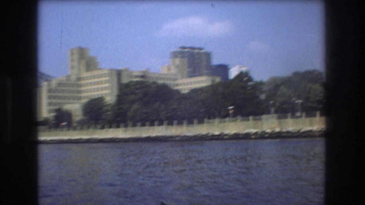 1975 Boston shoreline frame with river water in the foreground, a tree-lined embankment, and large pale city buildings softened by handheld 8mm motion.