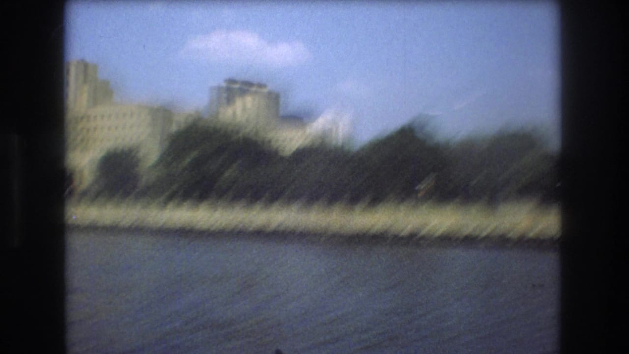 Motion-blurred 8mm frame from a boat showing Boston river water, a tree-lined shore, and softened city buildings in the background.