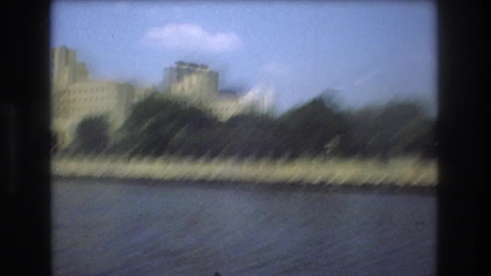 Motion-blurred 8mm frame from a boat showing Boston river water, a tree-lined shore, and softened city buildings in the background.