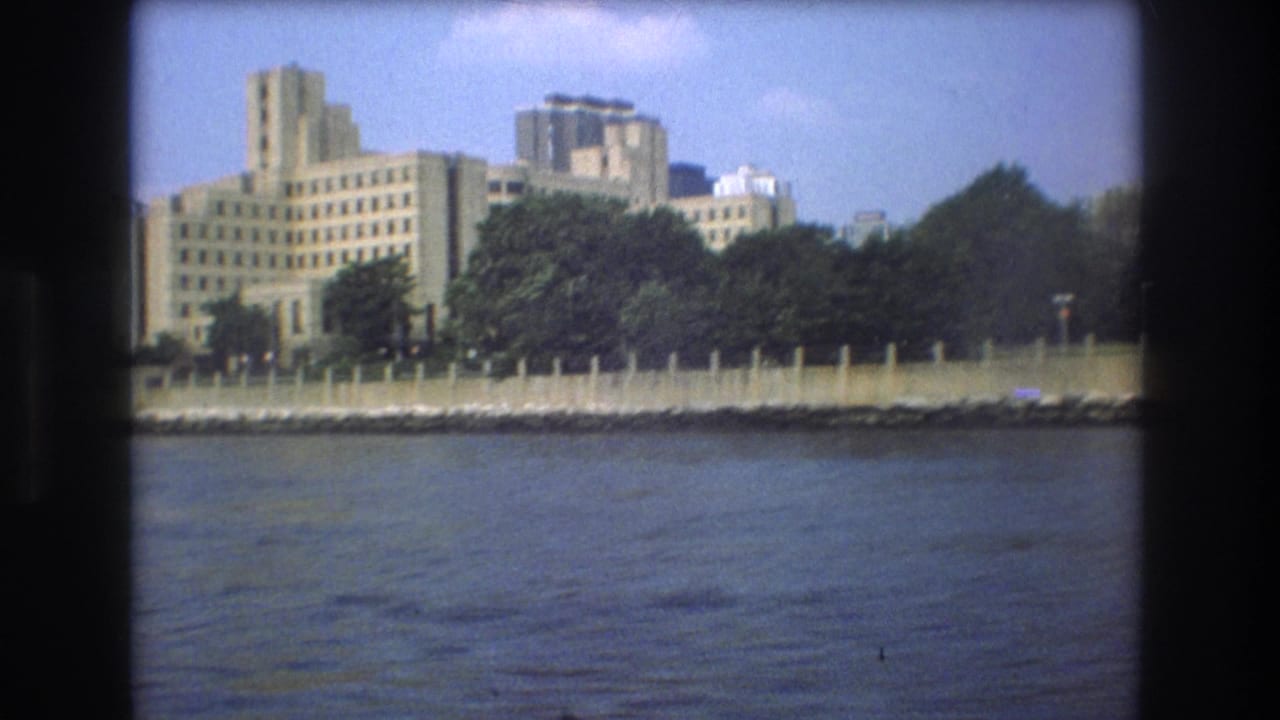 1975 Boston home-movie frame showing a large pale building with stepped towers behind a tree-lined shoreline across the water.