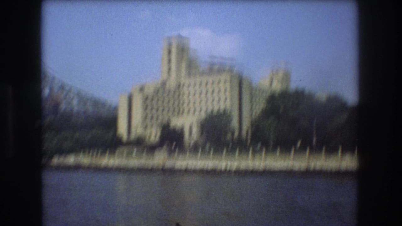 1975 Boston waterfront view with a pale stepped building, dark trees, low shore wall, and open water in the foreground.