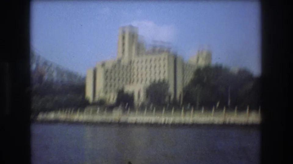 1975 Boston waterfront view with a pale stepped building, dark trees, low shore wall, and open water in the foreground.