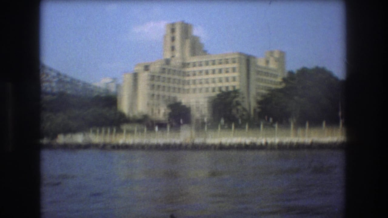 Boston 1975 home-movie frame showing the pale waterfront building across the water, with trees along the shore and a bridge structure partly visible at left.