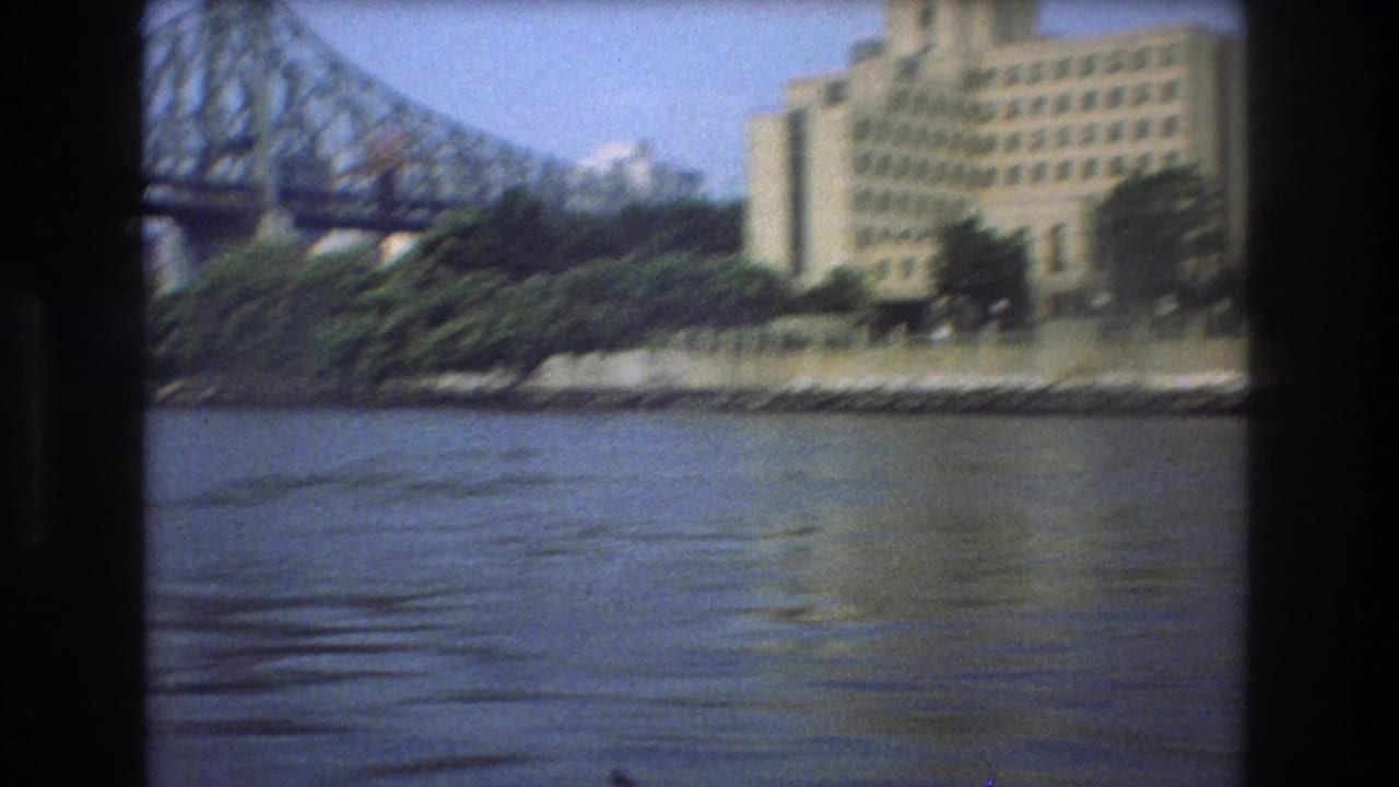 1975 Boston home-movie frame from a boat showing river water, part of a large steel bridge and a multi-story riverbank building in soft 8mm color.