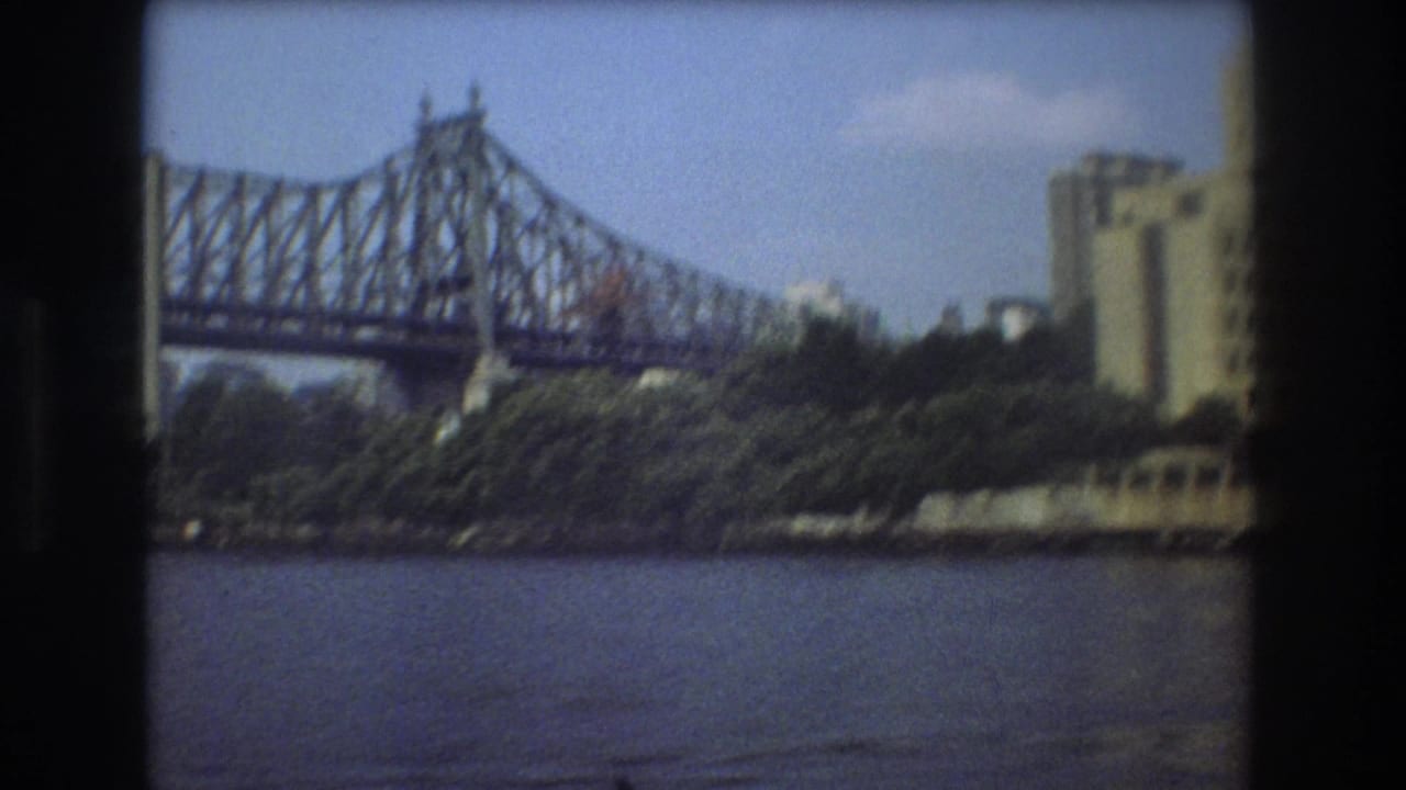 1975 Boston home-movie frame showing a steel truss bridge over a river, with trees and riverbank buildings visible beneath a blue sky.