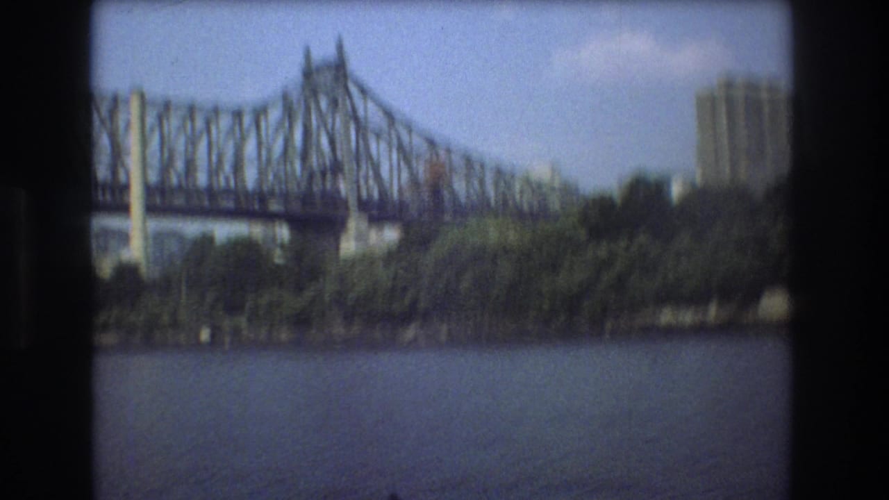 1975 Boston home-movie frame from a moving boat, with the bridge structure crossing above the river and a tall building partly visible beyond the trees.