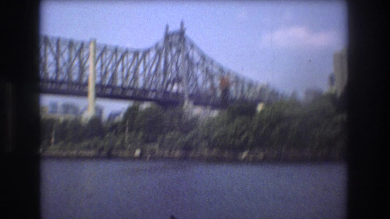 1975 Boston home-movie frame showing the bridge towers and trusswork over river water, with a tree-covered bank and light clouds in the sky.