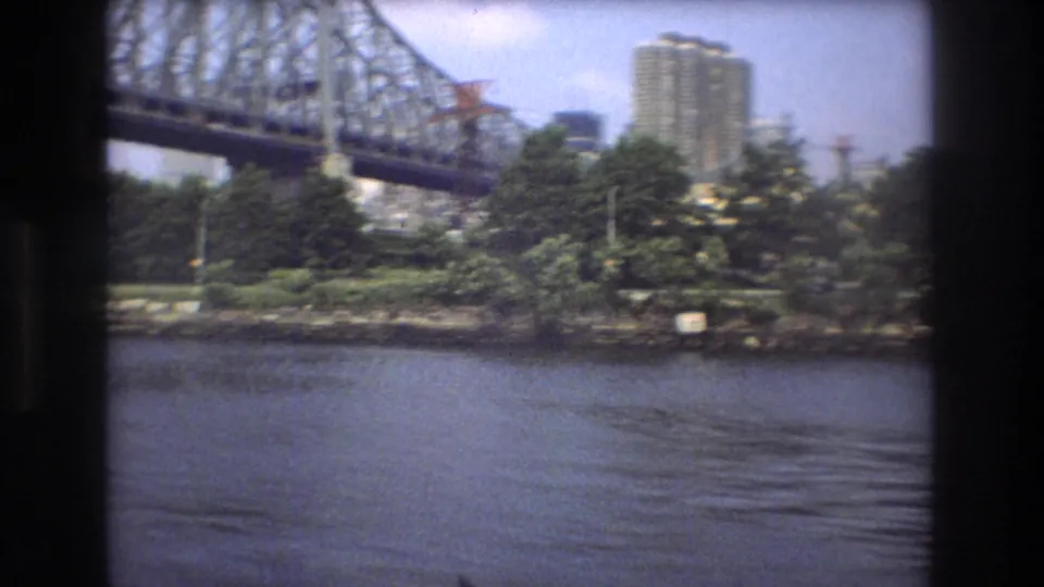1975 Boston home-movie frame of a steel bridge spanning the river, with waterfront trees and a distant high-rise building.