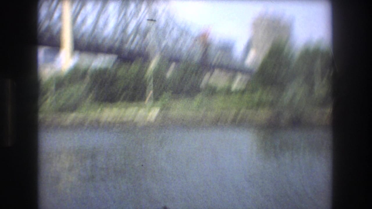 Motion-blurred 1975 Boston 8mm frame with a bridge, river, greenery, and distant building softened by camera movement.