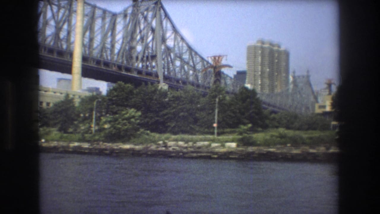 1975 Boston frame showing a large steel truss bridge from the river below, with trees, bridge supports, and distant high-rise buildings.