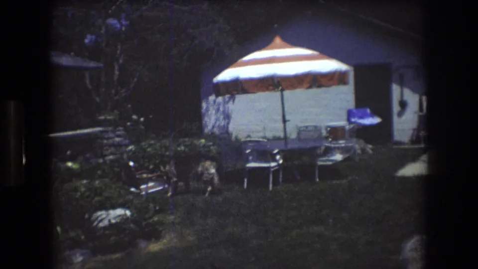 1976 Mt Bromley, Vermont 8mm frame showing a backyard with a red and white striped umbrella, metal chairs, a table, shrubs, and a dog moving across the lawn.