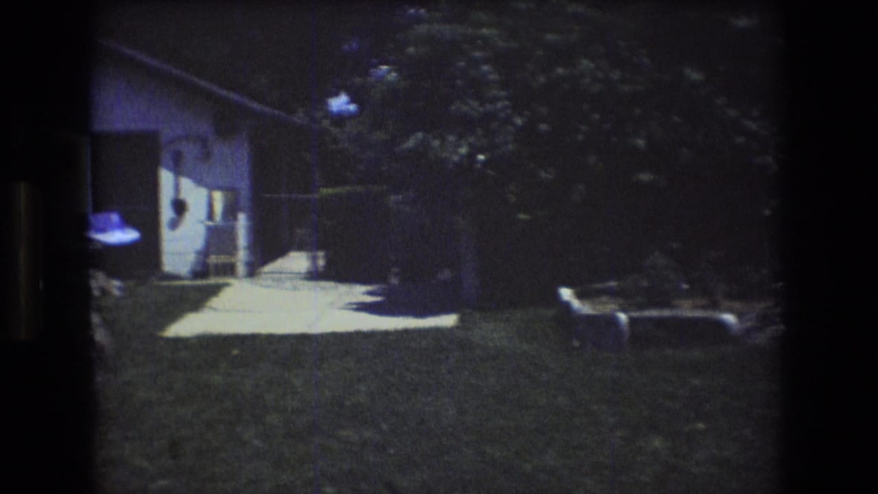 1976 Mt Bromley, Vermont backyard frame showing the pan moving toward shrubs and a concrete walkway, with part of the white-sided house and a dog near the right edge.