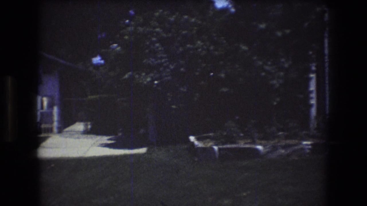1976 Mt Bromley, Vermont home-movie frame showing a shaded residential yard with a walkway, dense trees, lawn, and a low stone-edged planting bed.