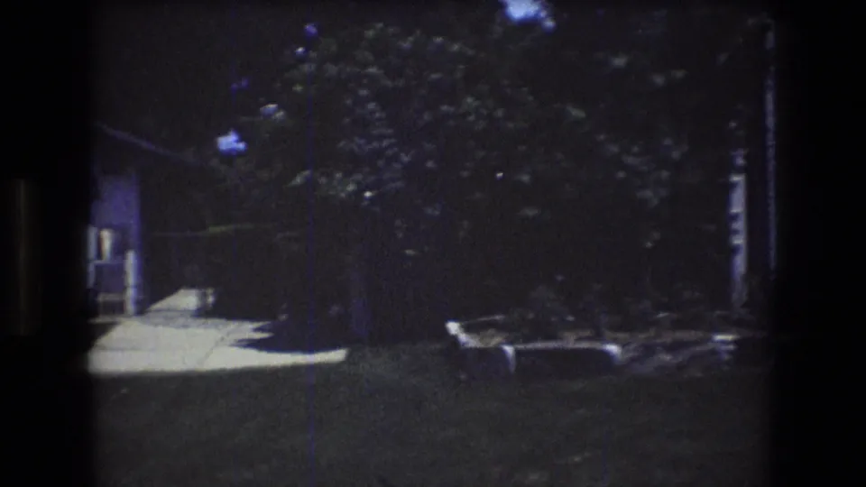 1976 Mt Bromley, Vermont home-movie frame showing a shaded residential yard with a walkway, dense trees, lawn, and a low stone-edged planting bed.
