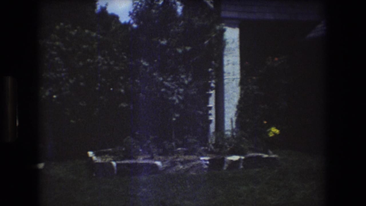 1976 Mt Bromley, Vermont frame showing a quiet house-side lawn with trees, shrubs, a white vertical exterior element, and a stone-edged planting bed.